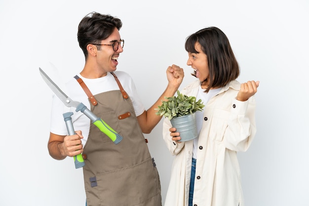 Jóvenes jardineros de raza mixta sosteniendo una planta y tijeras de podar aisladas sobre fondo blanco celebrando una victoria en la posición ganadora