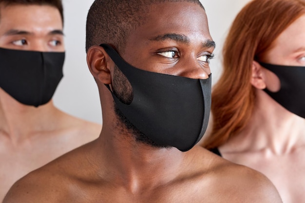 Jóvenes con máscaras médicas protectoras aisladas sobre fondo blanco en el estudio, hombres jóvenes sin camisa y mujeres pelirrojas mirando con confianza el concepto de corvid19 del coronavirus lateral enfocado en el hombre negro