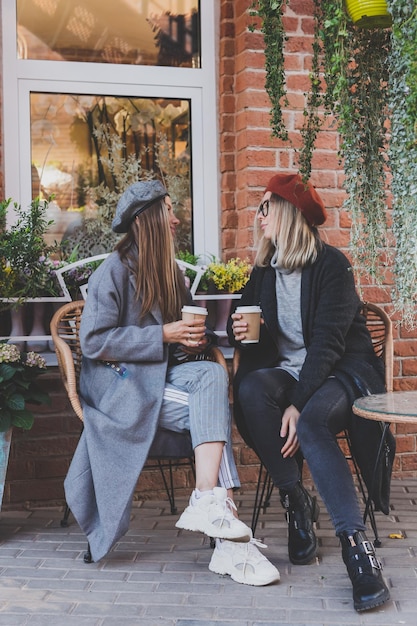 Jóvenes mejores amigas hablando entre ellas discutiendo temas interesantes durante el tiempo libre chicas hipster bromeando y divirtiéndose juntas disfrutando de la recreación con café para llevar en el fondo urbano