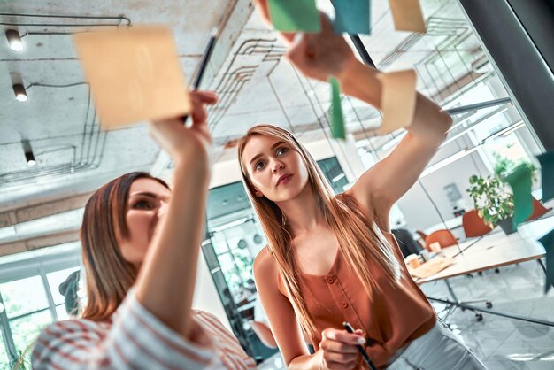 jóvenes modernos con ropa informal inteligente usando notas adhesivas mientras están de pie detrás de la pared de vidrio