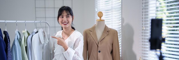 Foto jóvenes mujeres comerciantes asiáticas que usan su teléfono inteligente para grabar transmisión en vivo y presentar ropa nueva en el estante para vender en las redes sociales mientras trabajan y envían entregas para negocios de compras en línea