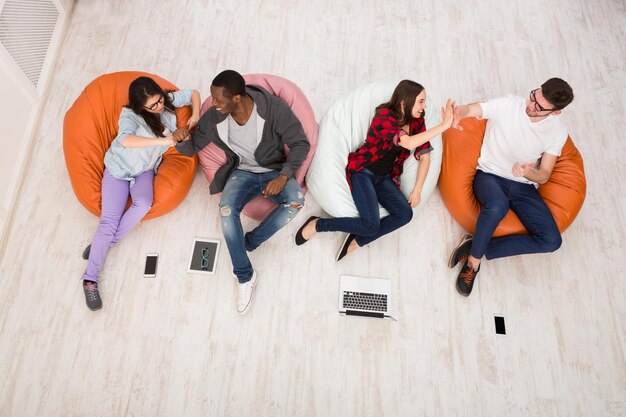 Los jóvenes multiétnicos, los estudiantes se relajan, se preparan para los exámenes en sillones puff, usando gadgets