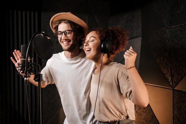 Jóvenes músicos atractivos cantando alegremente juntos grabando una nueva canción en un estudio de sonido moderno