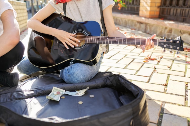 Jóvenes músicos callejeros, primer plano de la mano de una chica tocando guitarra acústica y estuche con dinero en efectivo en la acera