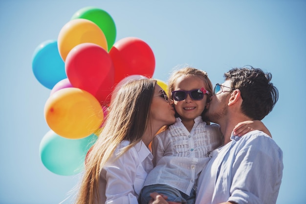 Jóvenes padres atractivos con globos besando a su pequeña hija en la mejilla