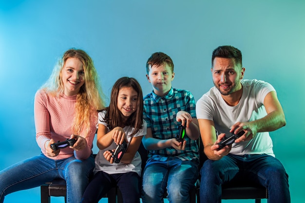 Jóvenes padres e hijos alegres jugando videojuegos juntos sobre una pared azul