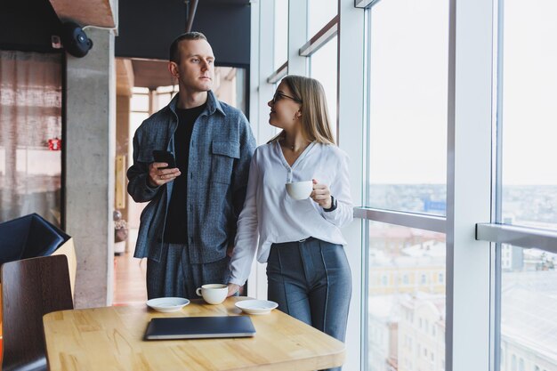 Jóvenes profesionales inteligentes con ropa informal revisando datos por teléfono y tomando notas mientras están de pie en una mesa en una oficina moderna cerca de una gran ventana en el último piso