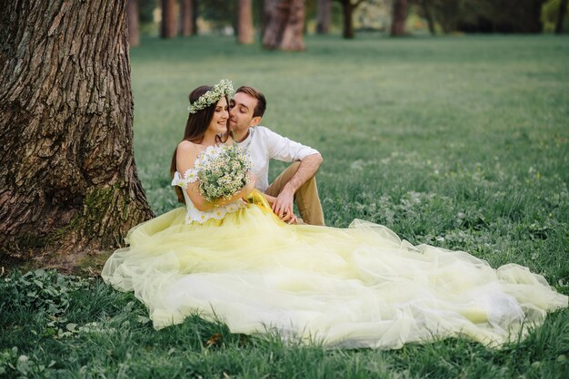 Jóvenes recién casados felices sentados en el césped cerca de un árbol en el parque