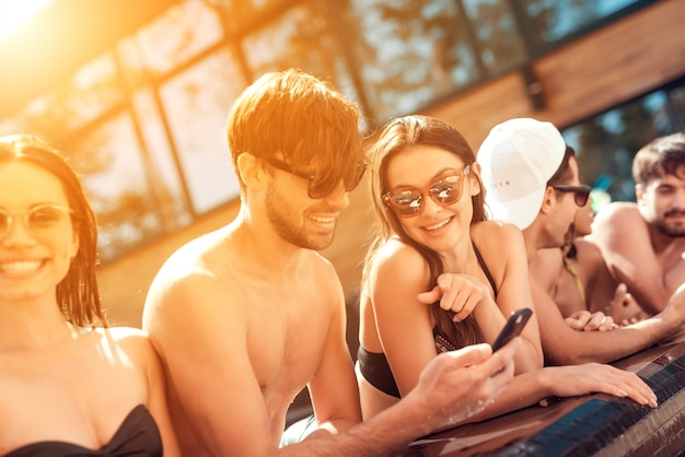 Jóvenes sonrientes amigos usando teléfono inteligente en la piscina