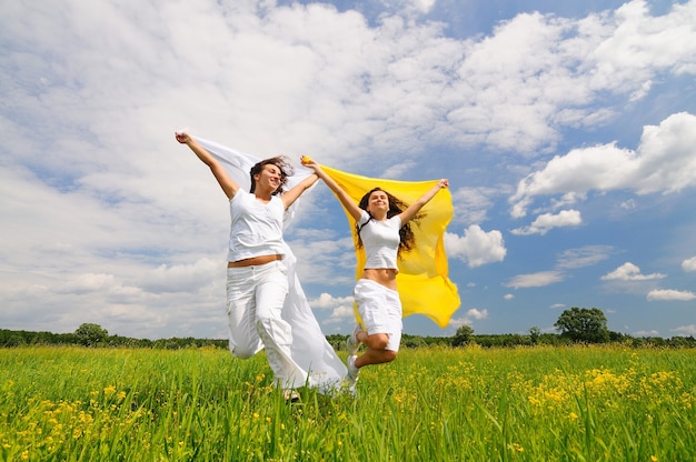 Jóvenes sonrientes a mujeres felices en ropa blanca saltando y sosteniendo chales de seda en manos levantadas en el prado y la hierba verde en verano en un día claro. Concepto de salud mental y energía positiva.