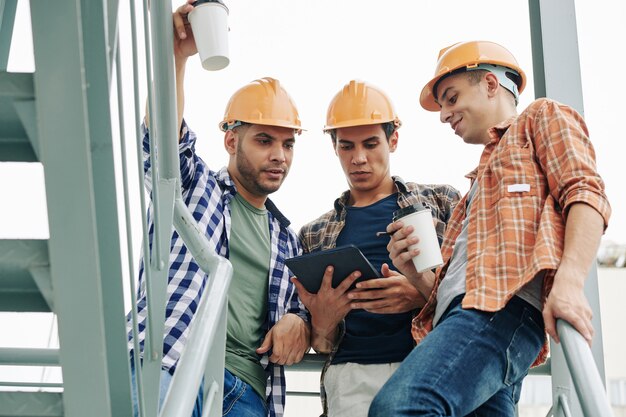 Jóvenes trabajadores de la construcción en cascos bebiendo café para llevar y revisando el correo electrónico del contratista en la tableta