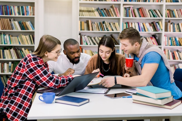 Jóvenes universitarios multiétnicos en la biblioteca que estudian juntos, están sonriendo, mirando el libro mientras encuentran nueva información útil para su examen