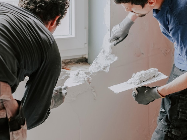 Jóvenes yeseros caucásicos aplican masilla con espátulas en la pared