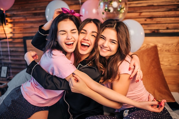 Jovens alegres e felizes abraçando uns aos outros. Eles se sentam juntos na cama e olham. Os adolescentes usam pijamas. Eles têm festa em casa.