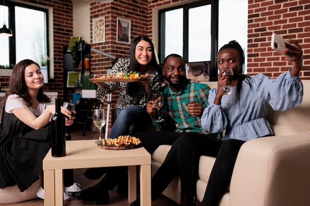 Foto jovens alegres multirraciais em casa comemorando aniversário enquanto tiram fotos de selfie juntos e bebem vinho. grupo de amigos bebendo bebidas alcoólicas enquanto se fotografam.