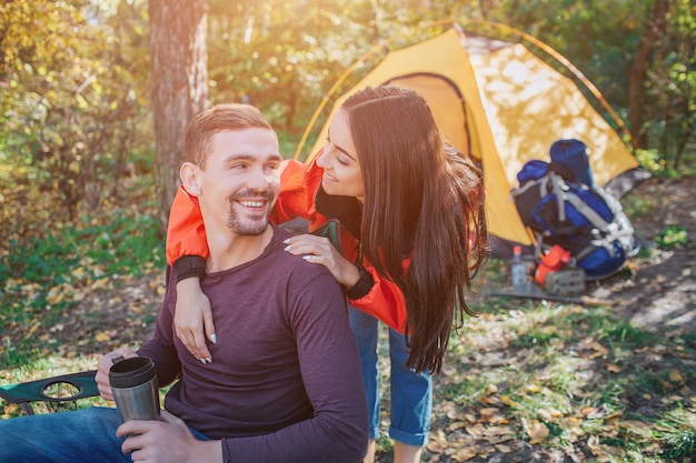 Jovens alegres se olham e sorriem. Ela está atrás dele. Jovem mulher abraça o homem. Ele é e segura uma garrafa térmica. Eles estão na floresta.