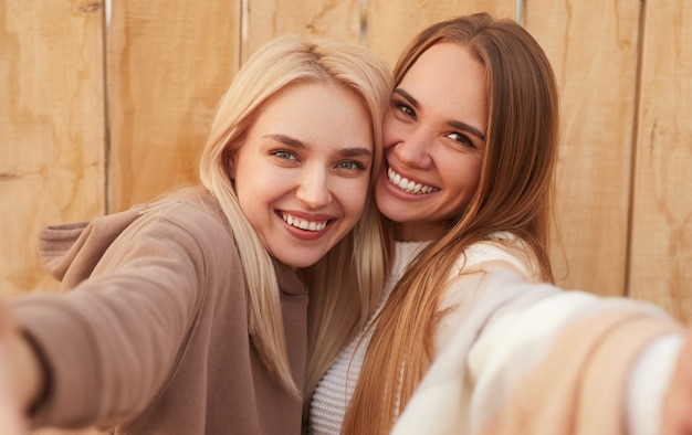 Jovens amigas alegres em roupas de malha quentes se abraçando e rindo enquanto tomam uma selfie juntas contra a parede de madeira