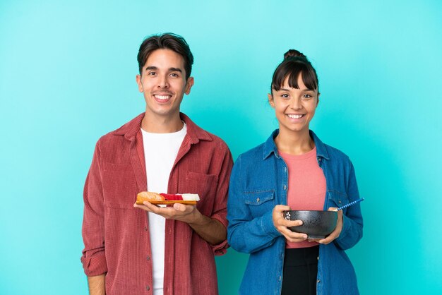 Jovens amigos de raça mista segurando sashimi e ramen isolados em fundo azul posando com braços no quadril e sorrindo