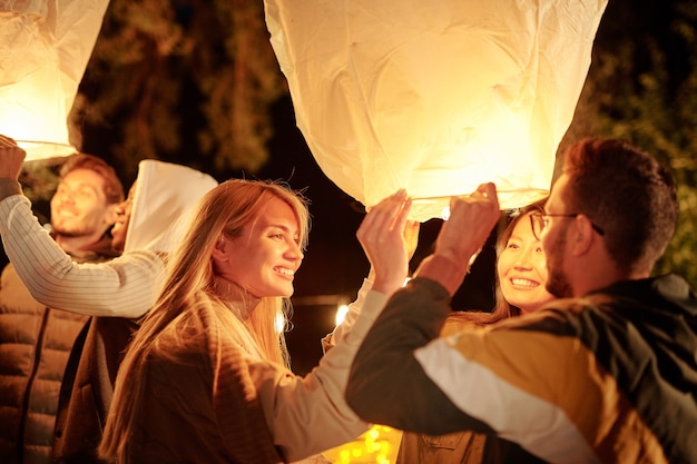 Jovens amigos interculturais sorridentes segurando grandes balões brancos iluminados enquanto aproveitam a festa noturna em ambiente natural