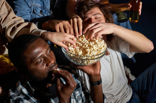 Jovens amigos multiétnicos assistem a filmes juntos em casa à noite, comendo lanches, vestidos casualmente, passando os fins de semana juntos