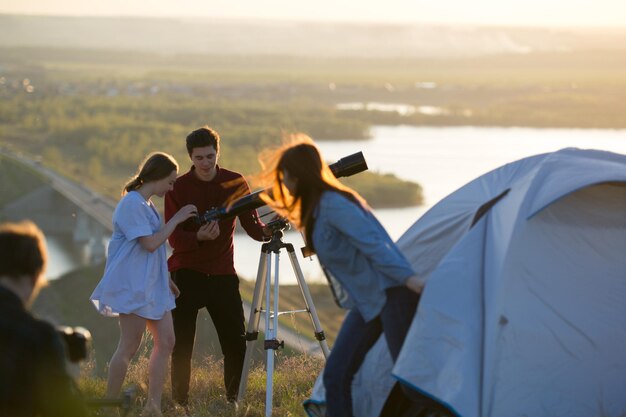 Jovens amigos olhando através do telescópio na colina em viajantes turísticos à noite de verão