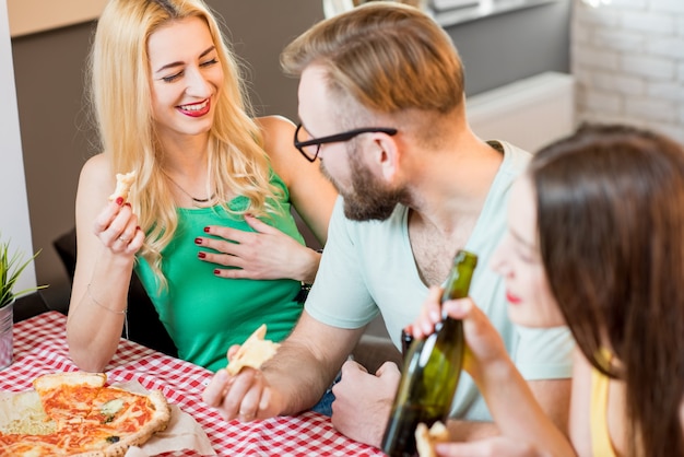 Jovens amigos vestidos casualmente com camisetas coloridas almoçando com pizza e cerveja em casa