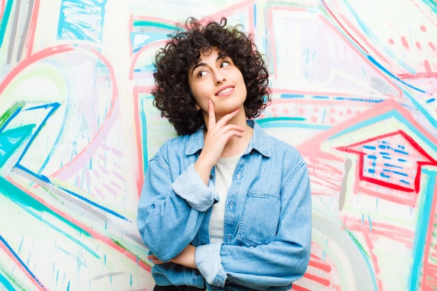 Foto jovens bonitas mulher afro sorrindo alegremente e sonhando acordado ou duvidando, olhando para o lado contra grafite