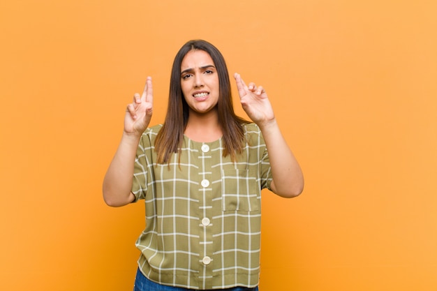 Jovens bonitas mulher cruzando os dedos ansiosamente e esperando a boa sorte com um olhar preocupado isolado contra a parede laranja