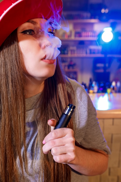 Jovens bonitas mulher de boné vermelho fumam um cigarro eletrônico na loja vape