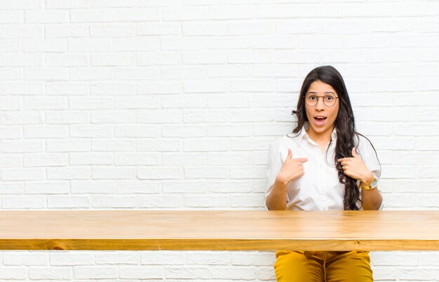 Jovens bonitas mulher latina se sentindo feliz, surpresa e orgulhosa, apontando para si mesma com um olhar animado e espantado, sentado na frente de uma mesa