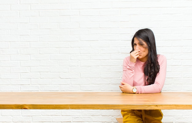 Jovens bonitas mulher latina sentindo nojo, segurando o nariz para evitar cheirar um fedor sujo e desagradável, sentado na frente de uma mesa