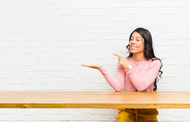 Jovens bonitas mulher Latina sorrindo alegremente e apontando para copyspace na palma da mão, mostrando ou anunciando um objeto sentado em frente a uma mesa