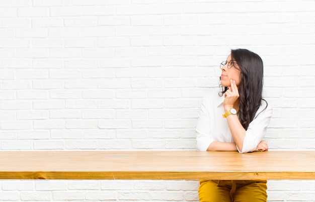 Jovens bonitas mulher Latina sorrindo alegremente e sonhando acordado ou duvidando, olhando para o lado sentado na frente de uma mesa