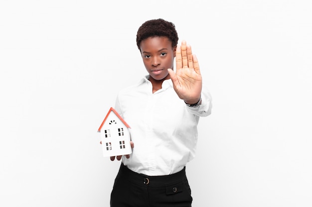 Jovens bonitas mulher negra olhando sério, popa, descontente e com raiva, mostrando a palma da mão aberta, fazendo o gesto de parada com um modelo de casa