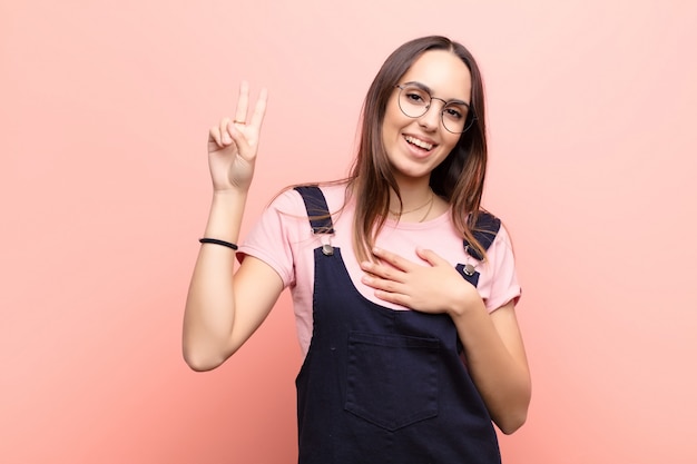 Jovens bonitas mulher olhando feliz, confiante e confiável, sorrindo e mostrando sinal de vitória, com uma atitude positiva contra a parede rosa