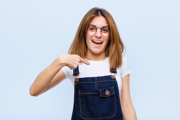 Jovens bonitas mulher olhando feliz, orgulhoso e surpreso, apontando alegremente para si mesmo, sentindo-se confiante e elevado sobre parede azul