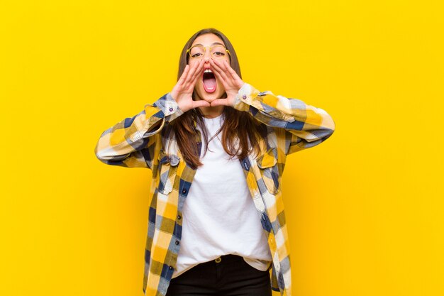 Jovens bonitas mulher se sentindo feliz, animada e positiva, dando um grande grito com as mãos ao lado da boca, gritando por cima do muro laranja