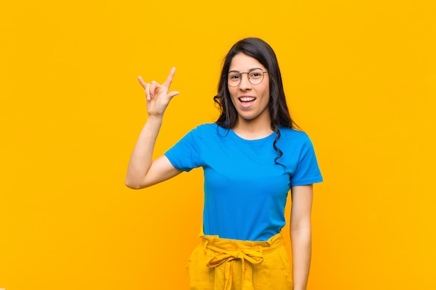 Jovens bonitas mulher sentindo feliz, divertido, confiante, positivo e rebelde, fazendo sinal de rock ou heavy metal com a mão contra a parede laranja