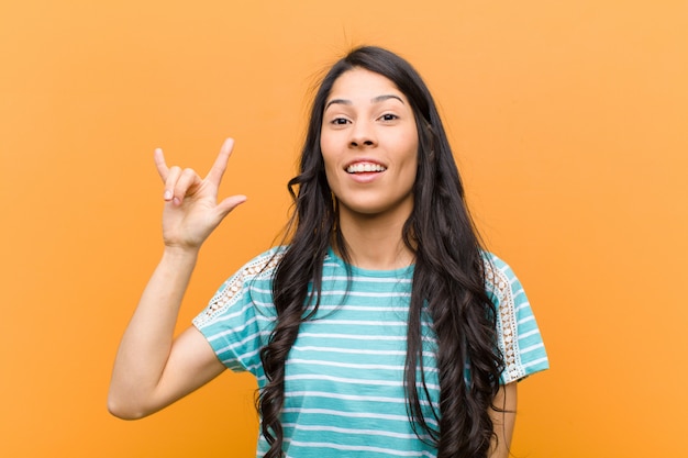 Jovens bonitas mulher sentindo feliz, divertido, confiante, positivo e rebelde, fazendo sinal de rock ou heavy metal com a mão contra a parede marrom