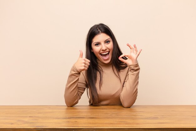 Jovens bonitas mulher sentindo feliz, espantado, satisfeito e surpreso, mostrando okey e polegares para cima gestos, sorrindo, sentado em uma mesa