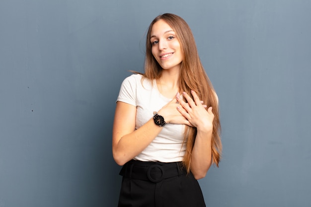 Jovens bonitas mulher sentindo romântico, feliz e apaixonado, sorrindo alegremente e segurando as mãos perto do coração contra a parede cinza