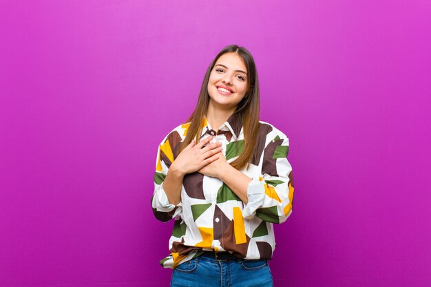 Jovens bonitas mulher sentindo romântico, feliz e apaixonado, sorrindo alegremente e segurando as mãos perto do coração contra a parede roxa