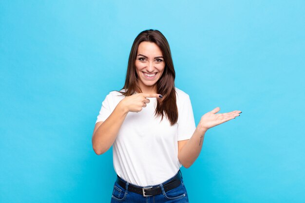 Foto jovens bonitas mulher sorrindo alegremente e apontando para copiar o espaço na palma da mão, mostrando ou anunciando um objeto contra a parede azul