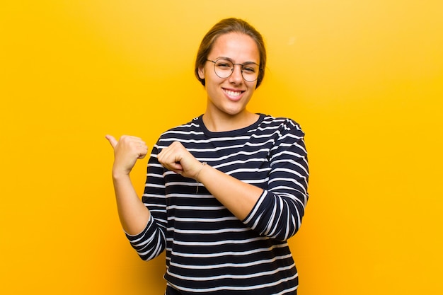 Jovens bonitas mulher sorrindo alegremente e casualmente apontando para copiar o espaço ao lado, sentindo-se feliz e satisfeito em laranja