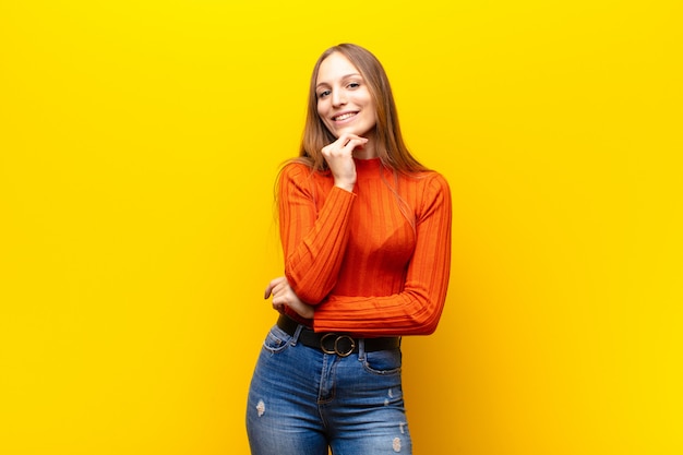 Jovens bonitas mulher sorrindo, curtindo a vida, sentindo-se feliz, amigável, satisfeito e despreocupado com a mão no queixo contra fundo laranja