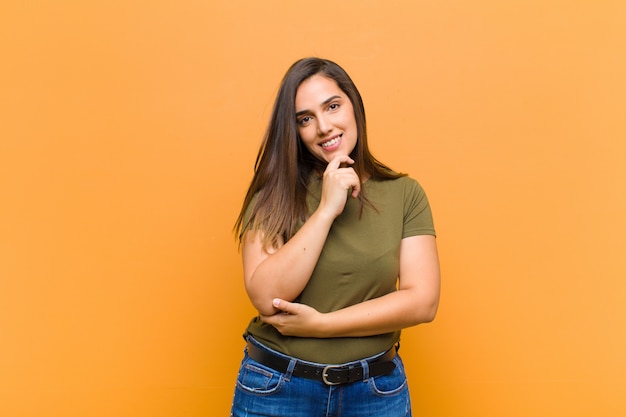 Jovens bonitas mulher sorrindo, curtindo a vida, sentindo-se feliz, amigável, satisfeito e despreocupado com a mão no queixo isolado contra a parede laranja