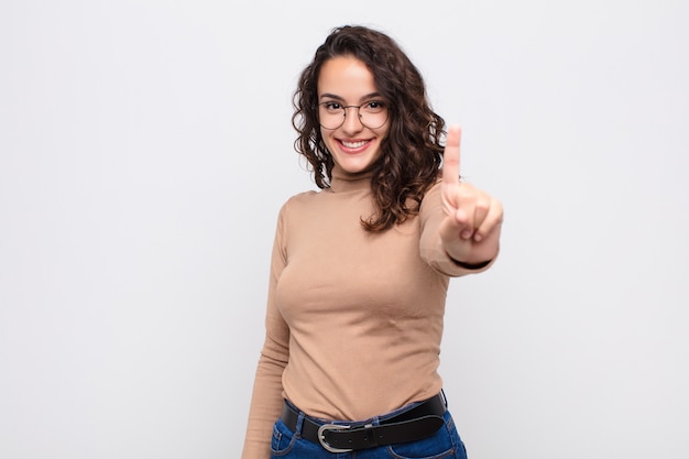 Jovens bonitas mulher sorrindo e olhando amigável, mostrando o número um ou primeiro com a mão para a frente, contando contra a parede branca