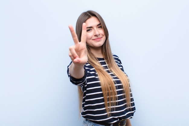 Jovens bonitas mulher sorrindo e olhando feliz, despreocupado e positivo, gesticulando vitória ou paz com uma mão contra a parede azul suave