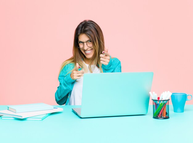 Jovens bonitas mulher trabalhando com um laptop sorrindo com uma atitude positiva, bem-sucedida e feliz, apontando para a câmera, fazendo sinal de arma com as mãos