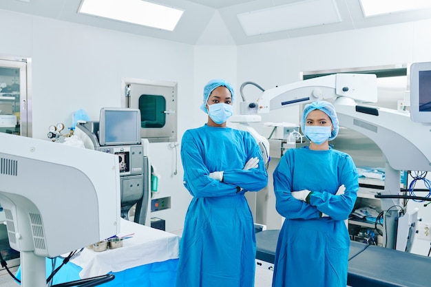 Jovens cirurgiões confiantes em aventais e máscaras médicas em pé na sala de cirurgia com os braços cruzados e olhando para a frente